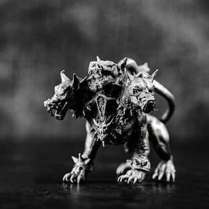 Puede incluir: Escultura en blanco y negro de Cerbero, el perro de tres cabezas de la mitología griega. La estatua presenta dientes afilados, collares con púas y una postura amenazante, con detalles intrincados y una estética oscura.