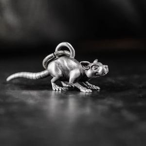 May include: A silver-colored rat pendant with a detailed design. The rat is depicted in a crouching position, with its tail extended. The pendant has a loop for attaching to a chain or keychain.