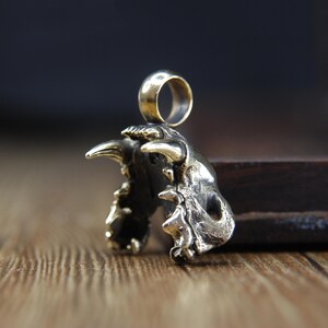May include: A detailed bronze pendant shaped like a stylized animal jaw with sharp teeth and a ring for a chain. The pendant has a weathered, antique finish, and is set against a blurred wooden background.