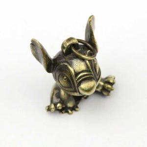 May include: A bronze-colored figurine of a cartoon character, featuring large ears, wide eyes, and a small body. The figurine has a loop at the top for attachment. The character is in a crawling position.