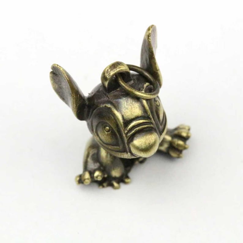 May include: A bronze-colored figurine of a cartoon character, featuring large ears, wide eyes, and a small body. The figurine has a loop at the top for attachment. The character is in a crawling position.