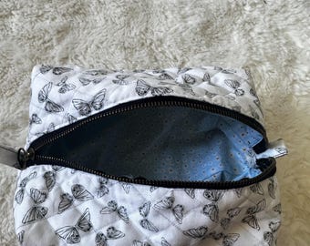 May include: White quilted cosmetic bag with a black butterfly pattern. The bag has a black zipper and a light blue interior with a small white dot pattern. A small gray strap is attached to the side.