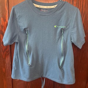 May include: A blue Champion t-shirt with two vertical zippers on the front. The shirt has a drawstring at the bottom and the Champion logo in green and yellow on the chest. The shirt is hanging on a white hanger.