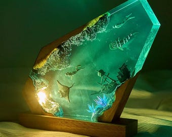 May include: A rectangular lamp featuring a wooden base and a translucent resin block illuminated with a green glow. The resin contains embedded miniature figures of a whale shark, a manta ray, a scuba diver, and a shipwreck within a sea scene.