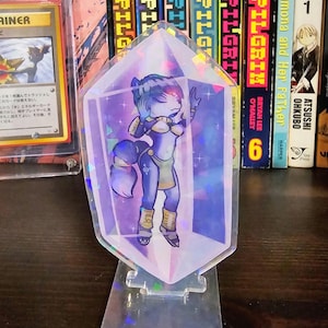Könnte beinhalten: Ein transparenter Acryl-Aufsteller mit einem holografischen, rautenförmigen Rahmen, der eine stilisierte Illustration einer violetten, anthropomorphen, fuchsgleichen Figur mit blauem Haar zeigt, die ein goldverziertes Outfit trägt.