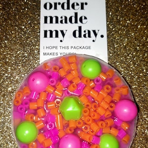 May include: A clear, circular container filled with orange, pink, and green beads. A white card with the text "Your order made my day" is next to it.