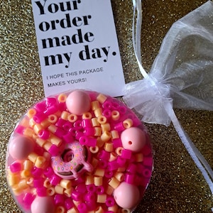 Op de afbeelding: Een helder, cirkelvormig harsstuk gevuld met roze en gele kralen en roze kralen. Een kleine donut-bedel staat in het midden. Een witte kaart met zwarte tekst luidt "Your order made my day". Een kleine witte mesh-tas is ook aanwezig.