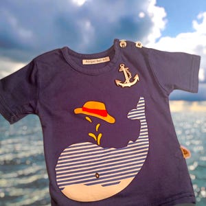 May include: Navy blue short-sleeved t-shirt with a whale design. The whale is white with blue stripes and a yellow and red hat. The shirt has an anchor and the text "Abigail Beli Art".
