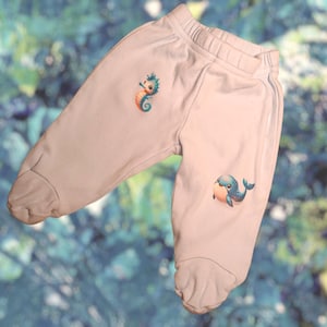 Puede incluir: Pantalones de bebé rosa claro con un diseño de caballito de mar y ballena. El caballito de mar es naranja y turquesa, la ballena es azul y melocotón. Los pantalones tienen cintura acanalada y pies.