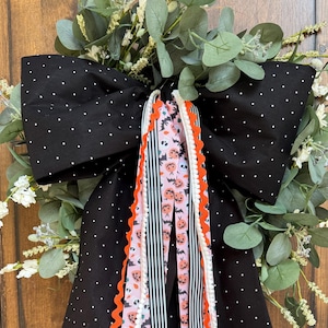 May include: A decorative wreath bow featuring a large black fabric base with white polka dots, layered with hanging ribbons in black and white stripes, orange rickrack trim, and a light pink pattern of pumpkins, ghosts, and bats.