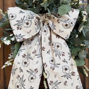 May include: A decorative wreath featuring green eucalyptus leaves and small white flowers, accented by a large, prominent fabric bow. The bow is made of cream-colored fabric with a repeating black and brown floral and botanical sketch pattern.