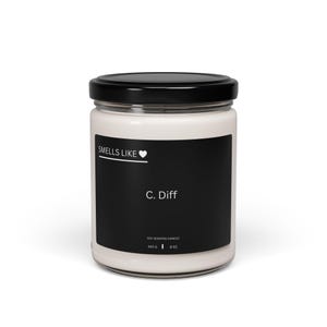 May include: A white soy scented candle in a clear glass jar with a black lid. The candle has a black label with the text "Smells Like" above a heart symbol, and "C. Diff" below. The label also indicates the candle weighs 340 g and 9 oz.