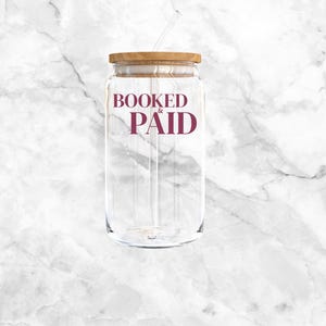 May include: Clear glass can-shaped tumbler with a wooden lid and a straw. The words "BOOKED & PAID" are printed in burgundy. The tumbler is designed for cold beverages.