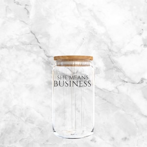 Può includere: Un bicchiere di vetro trasparente con coperchio in legno e cannuccia. Le parole "SHE MEANS BUSINESS" sono stampate in nero sul davanti. Sfondo bianco con motivo marmorizzato.