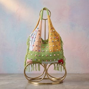 Könnte beinhalten: Eine bunte Patchwork-Tragetasche mit verschiedenen Texturen und Farben, darunter Grün, Gelb und Rosa. Die Tasche ist mit kleinen, bunten Perlen und Fransen verziert. Sie wird auf einem goldfarbenen Metallständer präsentiert.