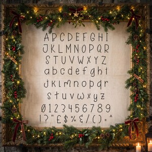 May include: A cross-stitch alphabet pattern on light fabric featuring uppercase and lowercase letters, numbers 0-9, and symbols. The pattern is framed by a decorative gold frame adorned with evergreen branches, red berries, and lights.