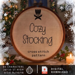 May include: A round wooden embroidery hoop containing beige fabric with black cross-stitched text that reads Cozy Stocking, a black icon above the text, and smaller black text below reading cross stitch pattern.