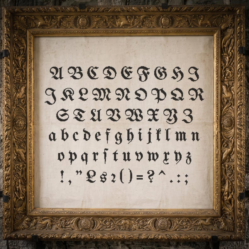 May include: A framed cross-stitch sampler on off-white fabric featuring the full alphabet in uppercase and lowercase letters, along with various punctuation marks in black thread, enclosed in an ornate, rectangular, antique gold-colored frame.