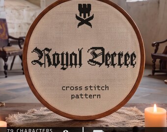Op de afbeelding: Een ronde houten borduurring met een kruissteekpatroon op gebroken wit stof. Het ontwerp bevat een zwart kroonicoon boven de tekst Royal Decree, gevolgd door de woorden cross stitch pattern, 79 characters, 33-38 st high, Aida 14/16/18.