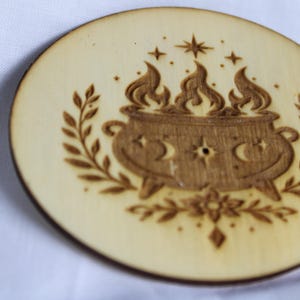 May include: A circular light-colored wooden coaster featuring a dark laser-engraved design of a three-legged cauldron with flames rising from the top. The cauldron is flanked by leafy branches and decorated with moon and star symbols.