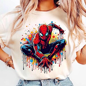 May include: A cream-colored t-shirt featuring a vibrant illustration of Spider-Man in a crouching pose. The superhero is depicted in his classic red and blue suit, with colorful paint splatter effects in the background.