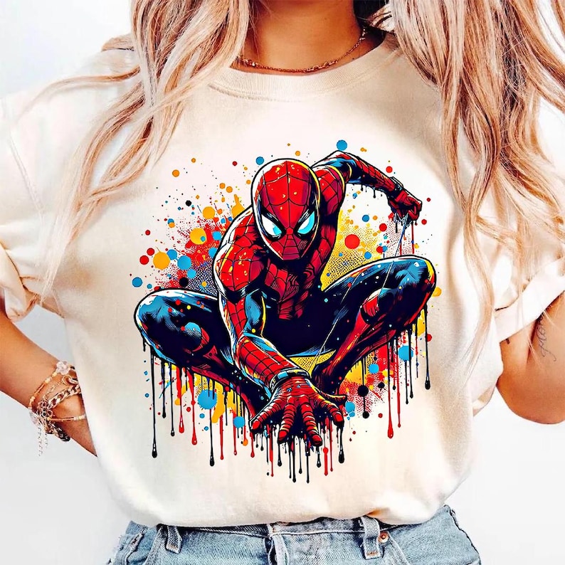 May include: A cream-colored t-shirt featuring a vibrant illustration of Spider-Man in a crouching pose. The superhero is depicted in his classic red and blue suit, with colorful paint splatter effects in the background.