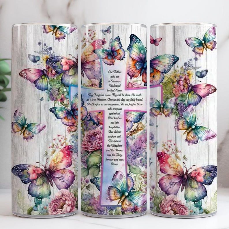 May include: Three tumblers with a floral and butterfly design. The tumblers feature a cross with the Lord's Prayer printed on it. The design includes colourful butterflies and flowers on a light wood grain background.