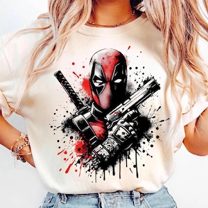 May include: Cream-colored t-shirt featuring a graphic of Deadpool, with a red and black color scheme. The design includes Deadpool's mask, a gun, and a katana, set against a backdrop of paint splatters.