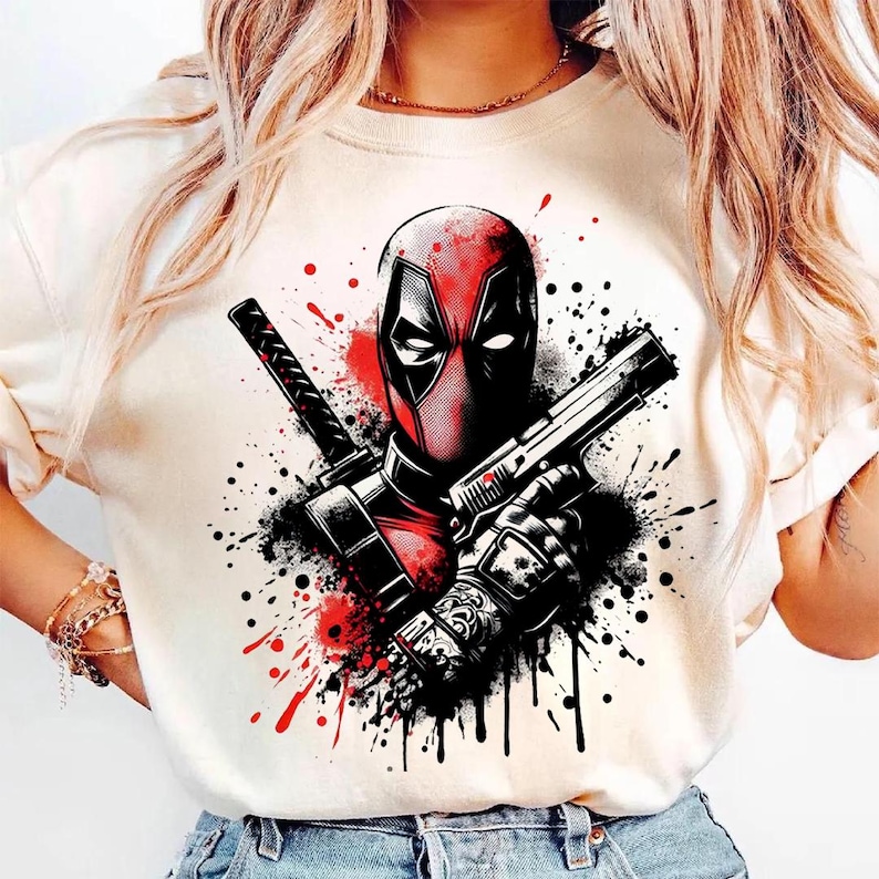 May include: Cream-colored t-shirt featuring a graphic of Deadpool, with a red and black color scheme. The design includes Deadpool's mask, a gun, and a katana, set against a backdrop of paint splatters.