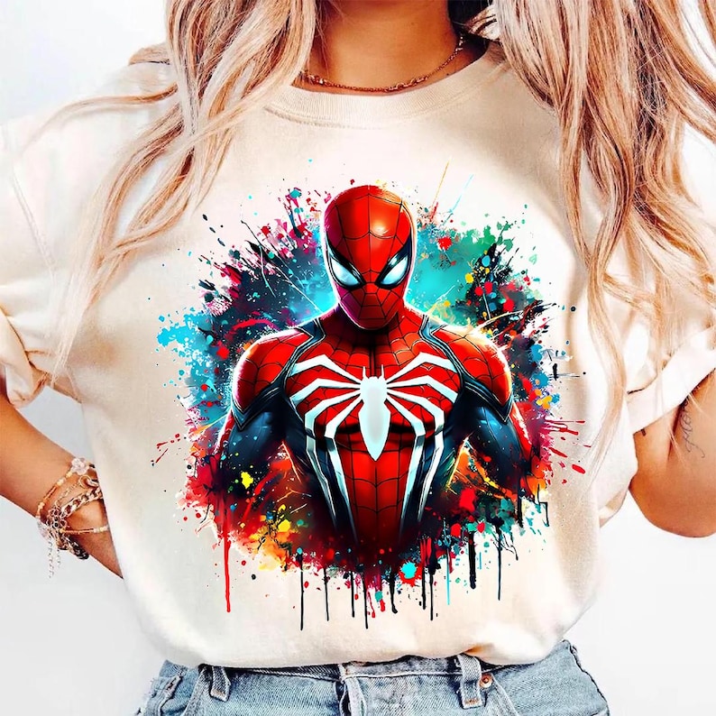 May include: A beige crew neck t-shirt featuring a vibrant graphic of Spider-Man. The superhero is depicted in his classic red and blue suit, set against a colorful splatter background. The design includes a white spider emblem on the chest.