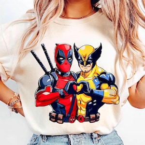 May include: Cream-colored t-shirt featuring a graphic of Deadpool and Wolverine forming a heart with their hands. Deadpool is in a red and black suit, and Wolverine is in a yellow and blue suit.