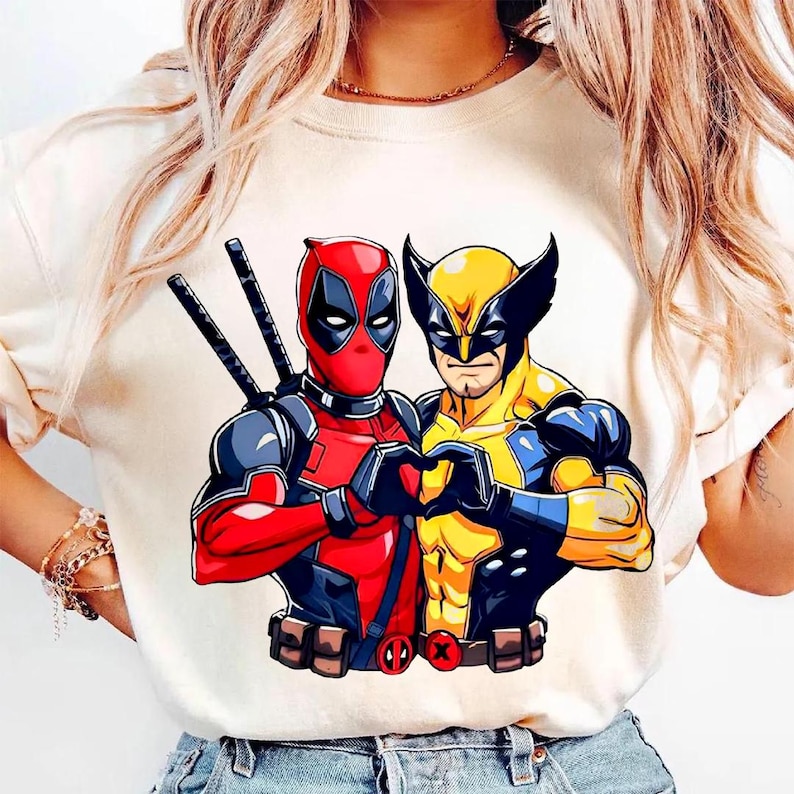 May include: Cream-colored t-shirt featuring a graphic of Deadpool and Wolverine forming a heart with their hands. Deadpool is in a red and black suit, and Wolverine is in a yellow and blue suit.