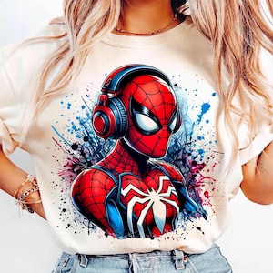 May include: Cream-colored t-shirt featuring a vibrant graphic of Spider-Man wearing headphones. The design showcases Spider-Man's iconic red and blue suit with a white spider emblem. The background includes colorful paint splatters.