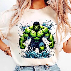 May include: Cream-colored t-shirt featuring a vibrant graphic of the Hulk, rendered in shades of green and blue. The Hulk is depicted in a dynamic pose, emerging from a splash of water. The design showcases the character's muscular physique and intense expression.