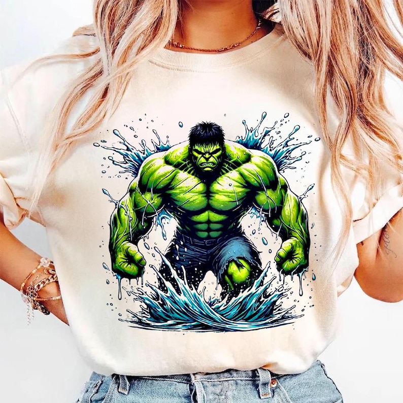 May include: Cream-colored t-shirt featuring a vibrant graphic of the Hulk, rendered in shades of green and blue. The Hulk is depicted in a dynamic pose, emerging from a splash of water. The design showcases the character's muscular physique and intense expression.