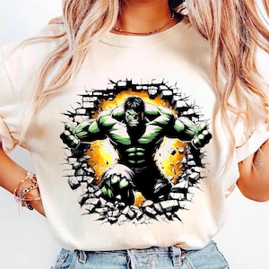 May include: Cream-colored t-shirt featuring a graphic of the Hulk bursting through a brick wall. The Hulk is depicted in green and black, with a yellow and orange background. The design is centered on the shirt.
