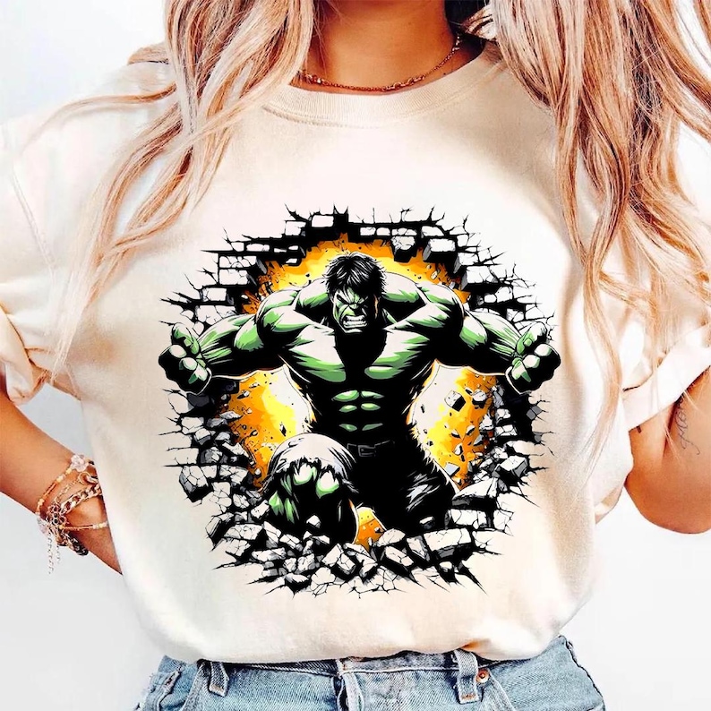 May include: Cream-colored t-shirt featuring a graphic of the Hulk bursting through a brick wall. The Hulk is depicted in green and black, with a yellow and orange background. The design is centered on the shirt.