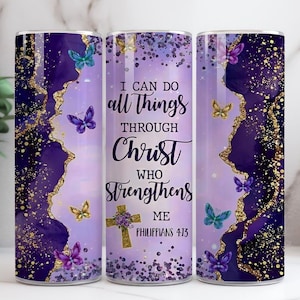 May include: A purple and gold tumbler with a design featuring butterflies and the text "I CAN DO all THINGS THROUGH CHRIST WHO STRENGTHENS ME PHILIPPIANS 4:13". The tumbler has a gold glitter design.