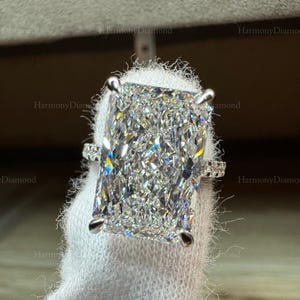 May include: A radiant-cut diamond ring. The large, rectangular-shaped diamond is the focal point, set with four prongs. The band features smaller accent diamonds. The ring is displayed on a white surface.