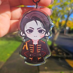 May include: A clear acrylic charm featuring a cartoon illustration of a character with dark hair and a black and red military-style jacket. The charm has a black cord for hanging. The character has a scar on their face.