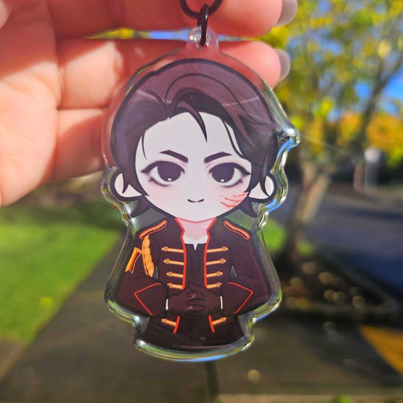 May include: A clear acrylic charm featuring a cartoon illustration of a character with dark hair and a black and red military-style jacket. The charm has a black cord for hanging. The character has a scar on their face.