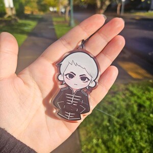 May include: A clear acrylic keychain featuring a cartoon character with white hair and a black uniform. The keychain is held in a hand against a blurred outdoor background.