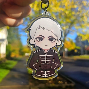 May include: A clear acrylic keychain featuring a cartoon illustration of a person with white hair and a stern expression. The person is wearing a dark brown military-style jacket with white accents. The keychain hangs from a black cord.
