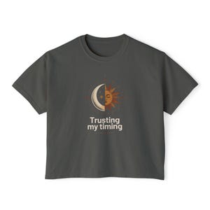 以下が含まれることがあります： ダークグレーの半袖Tシャツ。中央には放射状の線が描かれた太陽と月の顔が半分ずつ組み合わさったデザインがプリントされています。グラフィックの下には「Trusting my timing」という文字が、すっきりとした明るい色のサンセリフ体で記されています。