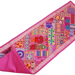 May include: A vibrant, rectangular patchwork table runner. The runner features a rich palette of pinks, reds, and other colors in a geometric pattern. The edges are trimmed with a dotted border. The underside is a solid pink fabric.