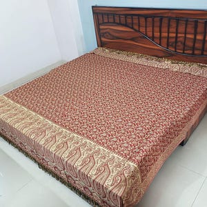 May include: A red and gold paisley patterned bedspread covers a bed. The bedspread has a detailed design with a border of large paisley shapes. The bedspread is on a wooden bed frame.