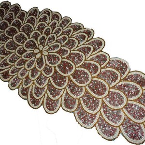 Puede incluir: Camino de mesa decorativo con diseño floral. El camino de mesa está hecho de cuentas marrones, blancas y rojas. El diseño se asemeja a una flor grande con múltiples pétalos. Los bordes están delineados en dorado.