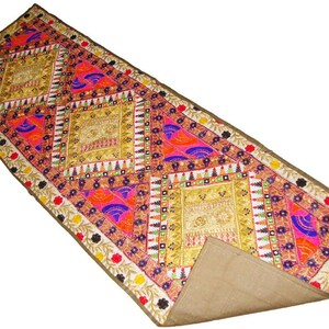 May include: A rectangular table runner featuring a vibrant patchwork design with gold, pink, orange, and blue embroidered patterns. The runner includes geometric diamond shapes, small mirrors, and a floral border on a tan fabric base with a folded corner.
