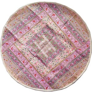May include: A round, floral-patterned textile in shades of pink, cream, and gold. The design features a central square surrounded by diagonal stripes with embroidered floral motifs. The edges are finished with a cream-colored border.