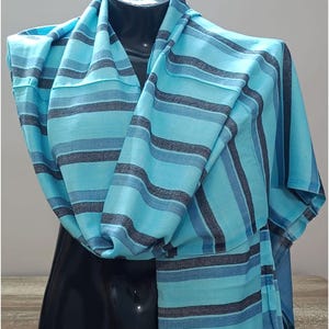 May include: A lightweight scarf featuring a horizontal striped pattern in shades of bright turquoise, medium blue, and dark charcoal grey. The fabric is draped in soft folds, displaying a consistent woven texture throughout the entire length of the piece.